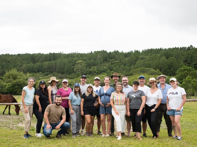 Foto de grupo dos participantes de uma edição, no campo, com os cavalos ao fundo.