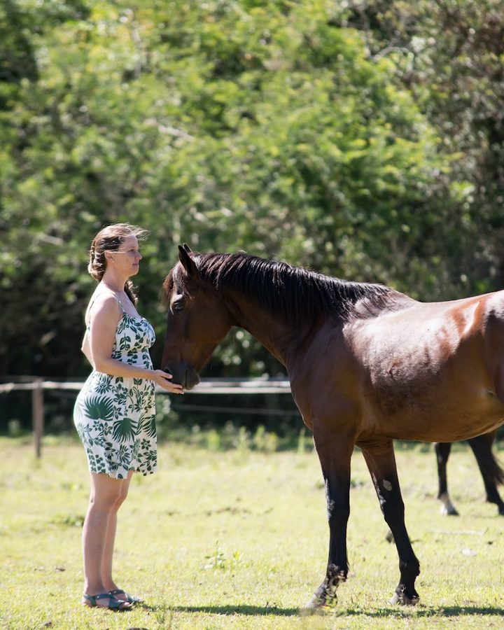 A Dra. Alice Prompt, de vestido florido, frente a frente com um cavalo baio, a mão no focinho dele.